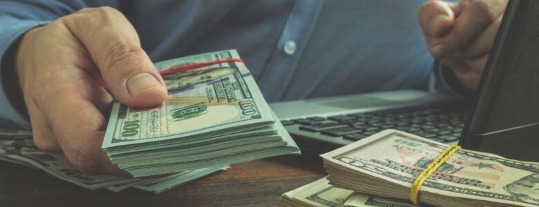 A person in a blue shirt sitting at a desk, holding a stack of U.S. hundred-dollar bills secured with a red band. Additional stacks of cash, secured with rubber bands, are placed on the wooden table next to a laptop keyboard.