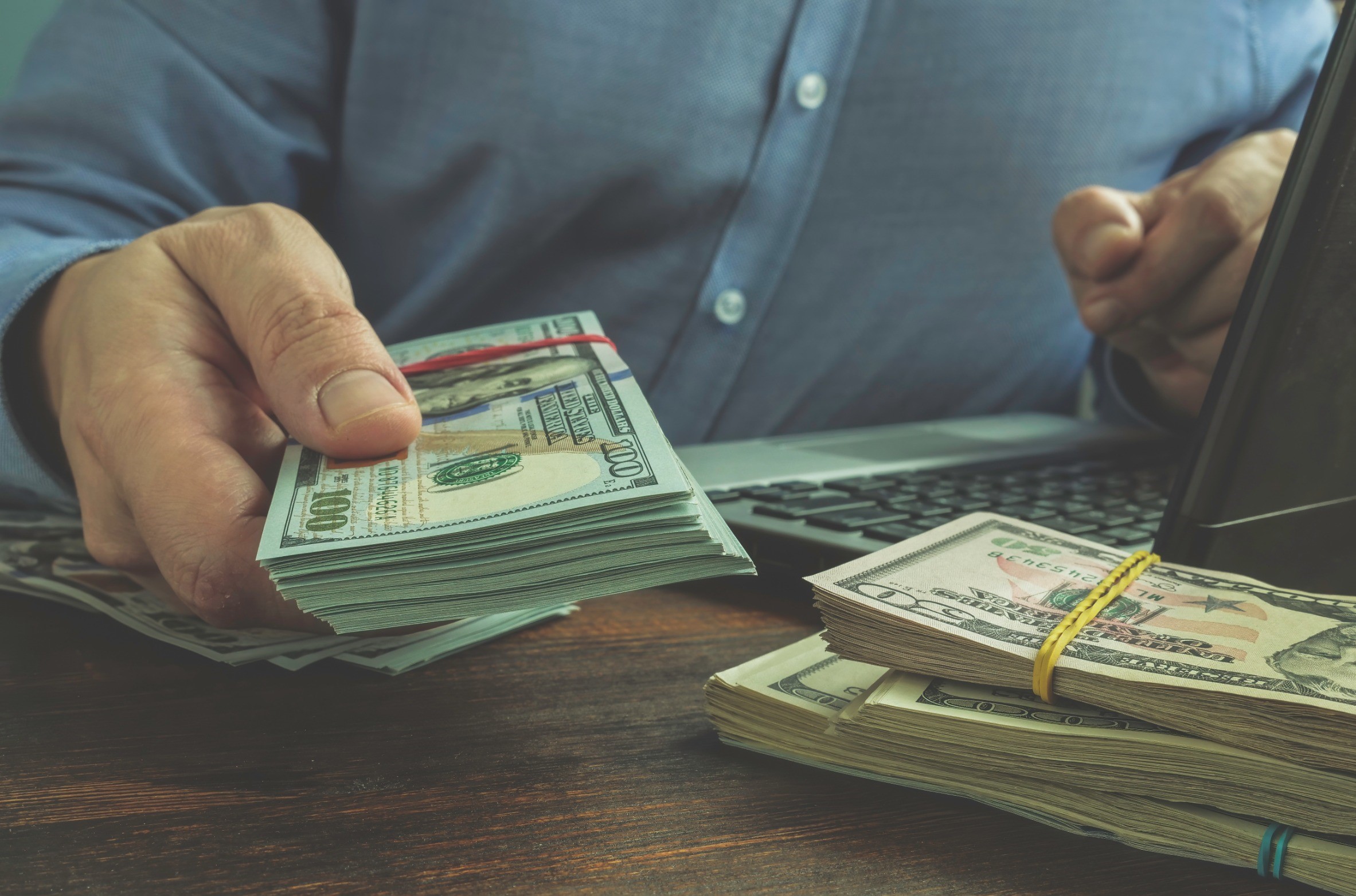 A person in a blue shirt sitting at a desk, holding a stack of U.S. hundred-dollar bills secured with a red band. Additional stacks of cash, secured with rubber bands, are placed on the wooden table next to a laptop keyboard.