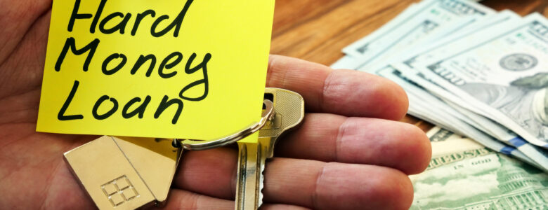 A close-up of a hand holding a yellow sticky note with "Hard Money Loan" written on it, along with house-shaped keychains and keys. In the background, there is a wooden desk with stacks of U.S. dollar bills, a calculator, and a closed notebook.