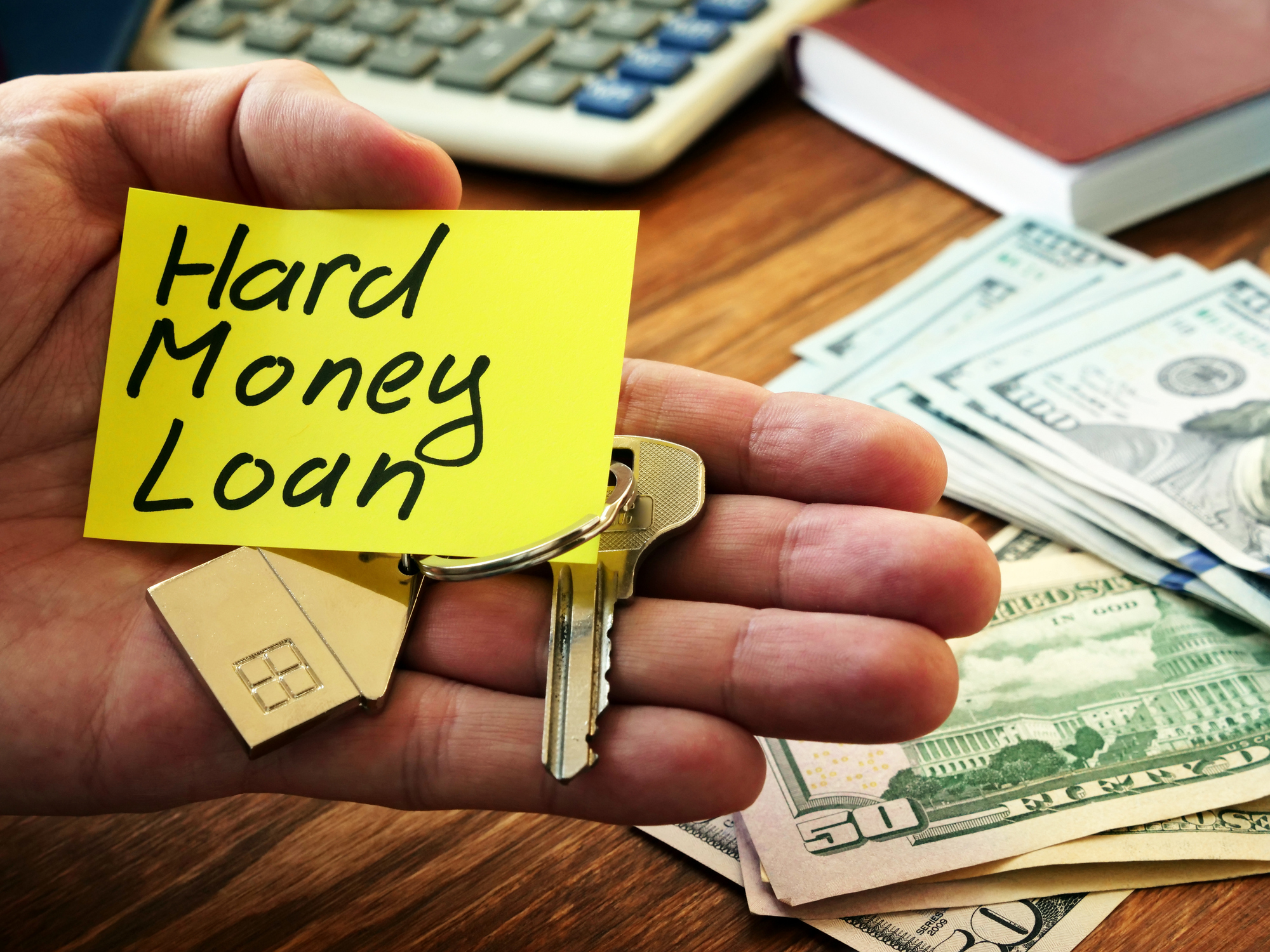 A close-up of a hand holding a yellow sticky note with "Hard Money Loan" written on it, along with house-shaped keychains and keys. In the background, there is a wooden desk with stacks of U.S. dollar bills, a calculator, and a closed notebook.