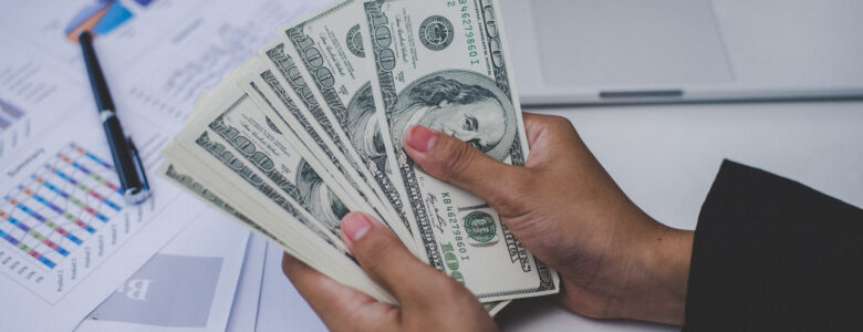 A person holding a stack of U.S. dollar bills over a desk, symbolizing financial transactions or budgeting. In the background, there is a laptop and printed charts with graphs and data, indicating financial planning, investment, or business activities.