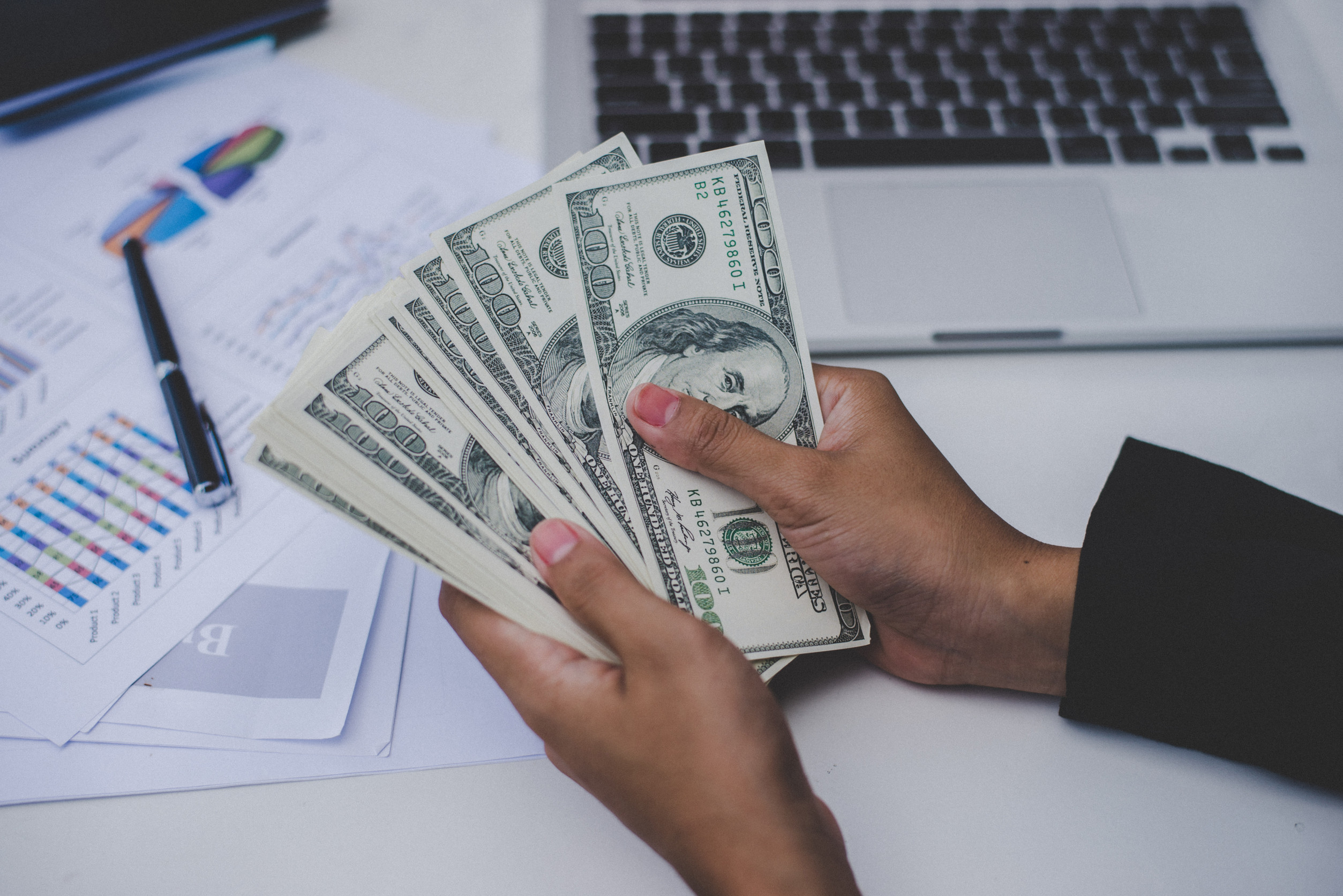 A person holding a stack of U.S. dollar bills over a desk, symbolizing financial transactions or budgeting. In the background, there is a laptop and printed charts with graphs and data, indicating financial planning, investment, or business activities.